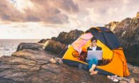 Girl working on laptop sitting in a tent on a cliff top